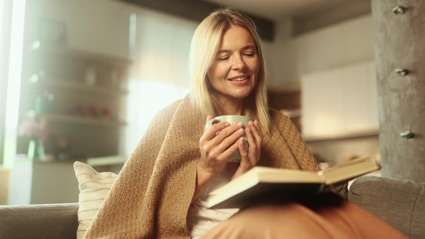 Portrait of charming blond mature woman under the blanket with cup of hot tea or coffee and reading book literature at home Calm relaxed female enjoying weekend leisure time alone indoors Royalty-Free Stock Footage #1106880421