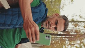 Vertical video, Mature man tourist wearing casual clothes sitting on bench scrolling smartphone