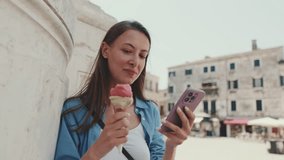 Close up, beautiful girl with long brown hair, dressed in blue shirt, enjoys ice cream and looks through photos, videos on mobile phone while standing on square of old city
