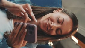Vertical video, Cute smiling girl with long brown hair, dressed in blue shirt, while traveling by suburban train, scrolls on smartphone screen