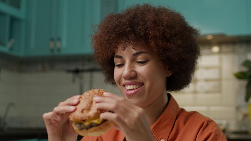 African American Woman Eating Burger, Close Up. Hungry black female biting cheeseburger at home. Royalty-Free Stock Footage #1106889501
