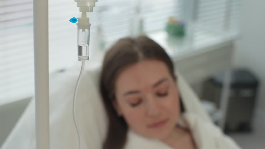 Close-up high-angle view of sick young woman lying in bed at clinic. Ill female patient with intravenous drip at hospital ward. Pretty lady receiving medication via intravenous therapy, slow motion. Royalty-Free Stock Footage #1106893621