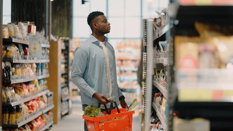 African American man with basket in hand chooses product in grocery store. Concept of shopping and shelves with organic food in supermarket slow motion 스톡 비디오