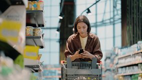 Woman picks up product from shelf pushing shopping cart in light supermarket. Brunette compares products with smartphone information. Shopping at grocery store - Powered by Shutterstock - Get 15% off with code: PIKWIZARD15