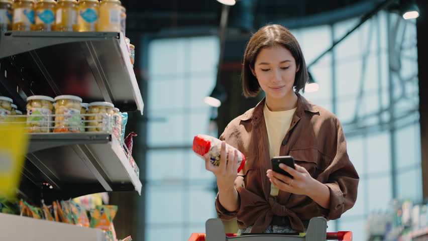 Woman looks at list on mobile phone taking product from supermarket shelf. Lady stands in grocery store with cart. Shopping and choice of goods