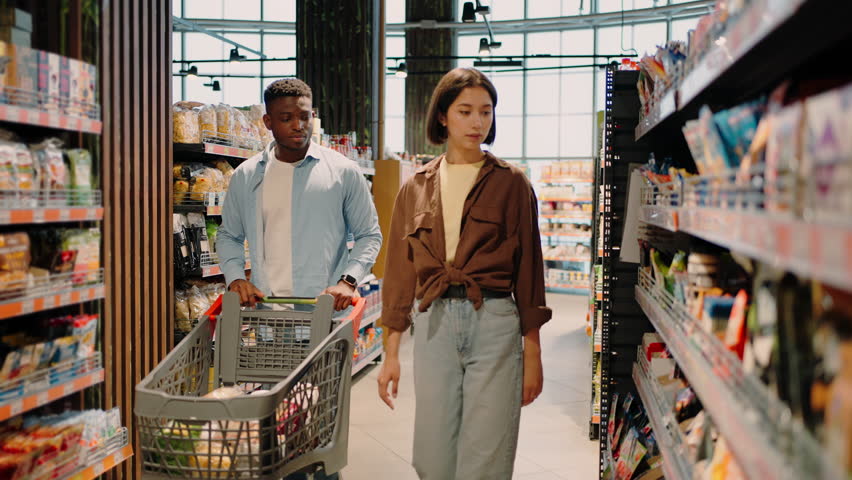 Couple of African American man and European lady choose food can by checking through smartphone. Choice of perfect goods on grocery store shelves together Royalty-Free Stock Footage #1106898381