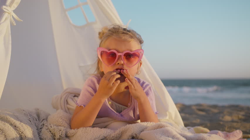 Hilarious preschooler kid playing, pretending glamorous lady in fashion heart shaped pink sunglasses 4K. Funny little girl expresses gestures and making funny faces while eating fresh juicy cherry Royalty-Free Stock Footage #1106899153