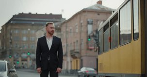 Businessman in formal suit standing in the central street with traffic. Looking confident and serious. Man with beard and mustache standing by the tram and cars driving through the town. 