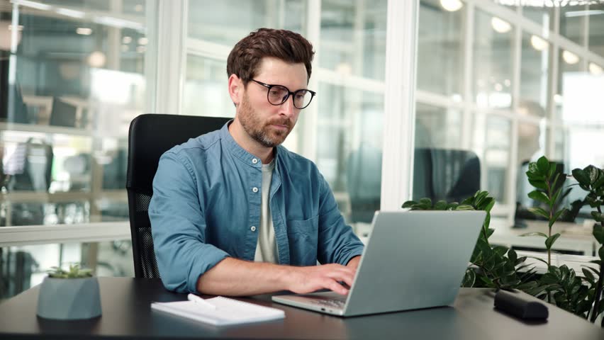 Concentrated businessman in glasses making notes in notepad using laptop. Young man typing on keyboard doing paper work makes online reporting of company at office place. Remote job business concept.