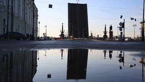 Open bascule bridge reflects on puddle surface at sunset in Saint Petersburg city. Cars drives on road. People walks on sidewalk in foreground. Selective focus. Real time video. Travel in Russia theme