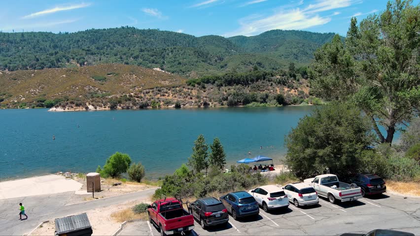 aerial footage of the rippling blue waters of Silverwood Lake with a beach and people swimming in the water and lush green trees, grass and plants, blue sky and clouds in Hesperia California USA