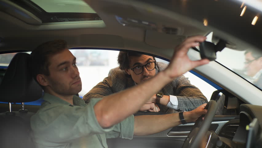 View through the side window of the car to stylish sales manager wearing business suit talking to male client sitting in car in showroom choosing automobile holding steering wheel, slow motion.