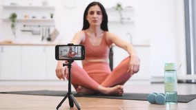 Blurred background of beautiful female meditator doing padmasana exercise in lotus pose while relaxing on fitness mat. Yong woman recording video while practicing yoga in bright stylish kitchen.