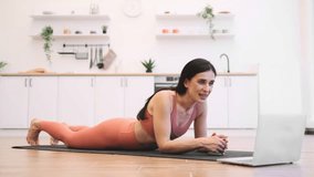 Strong female person standing in plank position and watching motivational video with online training on remote gadget. Flexible lady strengthening core and abs muscles while exercising on rug indoors.
