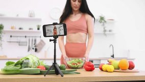 Stylish smartphone on stand capturing happy female in sports clothes showing bowl with nutritious dish. Attractive brunette lady sharing joy and pleasure of cooked meal with subscribers.