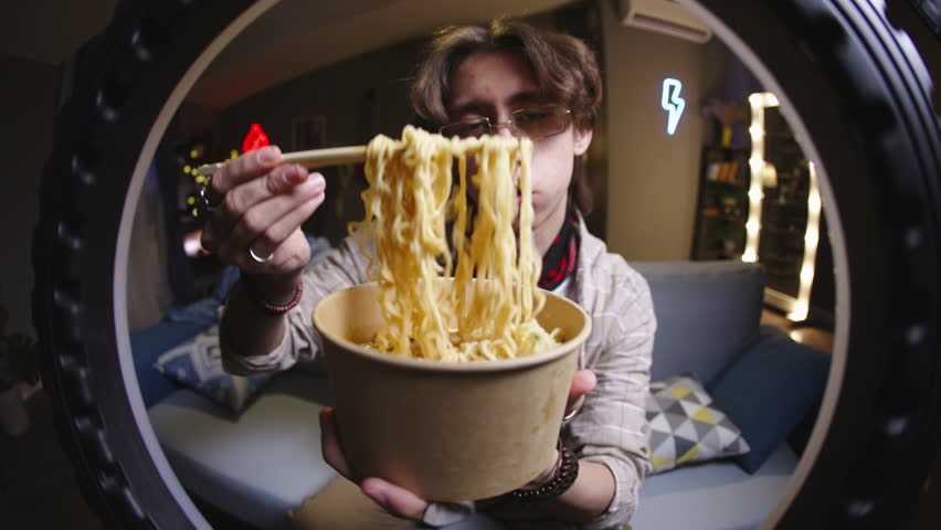 Asian guy with glasses shoots video while sitting on sofa in front of ring lamp how he eats noodles. Mookbang. Asian vlogger influencer social network, happy popular blogger. Concept of virtual life