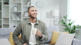 Happy young adult man drinking hot drink from mug. Satisfied smiling guy sipping tea sitting in cozy sofa in living room at home. indoor Mixed race male enjoys a beverage coffee cup or medicine - Powered by Shutterstock - Get 15% off with code: PIKWIZARD15