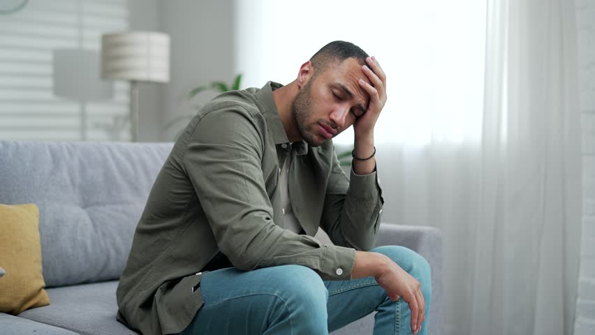 Silhouette of a young adult man suffering from depression sitting at home on the couch. The male has mental health issues upset guy alone near a window One anxious depressed man in despair indoor