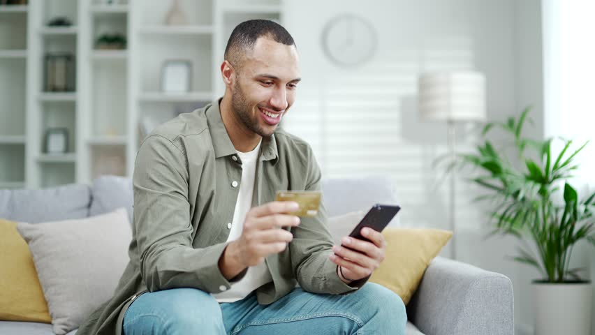 Young happy african american man sitting on the couch, using smart cell phone for online shopping with credit card on living room Indoor. Satisfied male orders or booking in store or paying bills Royalty-Free Stock Footage #1106930601