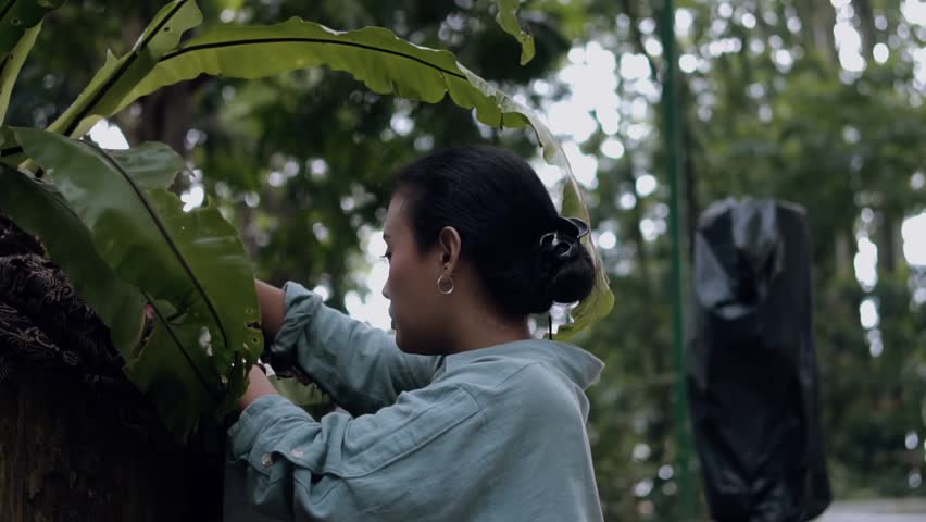 Profile view of a young woman in a park taking a red traditional Indonesian mask (Topeng) from concealment, looking inside it, and beginning to place it over her face