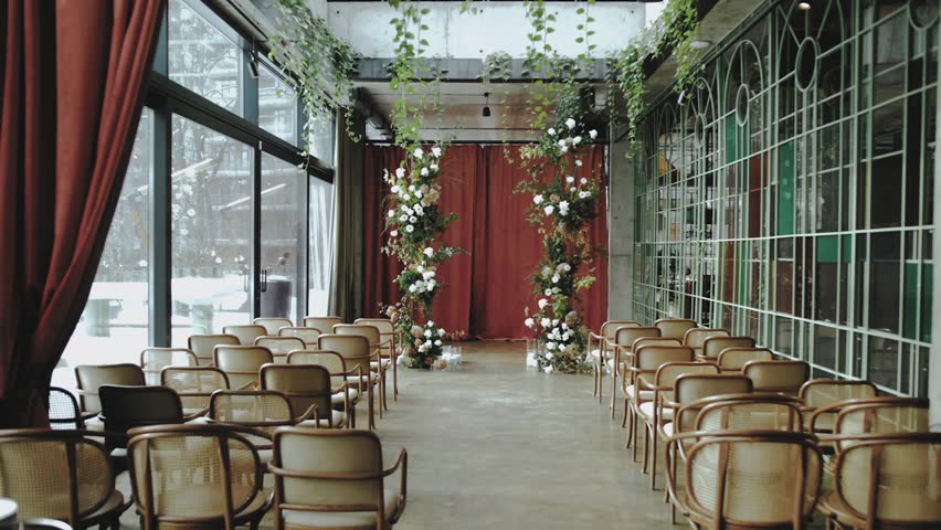 Wedding arch decorated with pastel faded flowers and cream-colored wooden chairs, indoor wedding celebration. Rustic wedding, dolly shot.