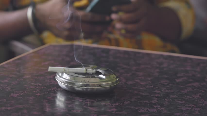 A closeup of a burning, smoky cigarette rests on a silver ashtray on the table, while a man browses his mobile in the background inside the house.smoking kills.quit smoking concept Royalty-Free Stock Footage #1106949175