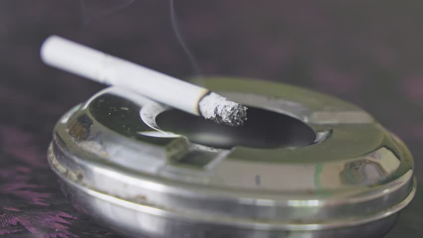 A closeup of a burning, smoky cigarette rests on a silver ashtray on the table inside the house.smoking kills.quit smoking concept Royalty-Free Stock Footage #1106949177