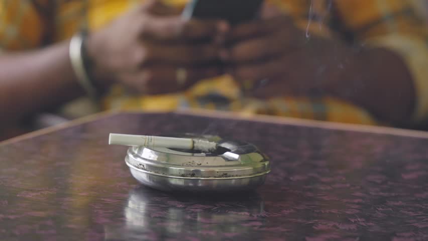A closeup of a burning, smoky cigarette rests on a silver ashtray on the table, while a man browses his mobile in the background inside the house.smoking kills.quit smoking concept Royalty-Free Stock Footage #1106949179