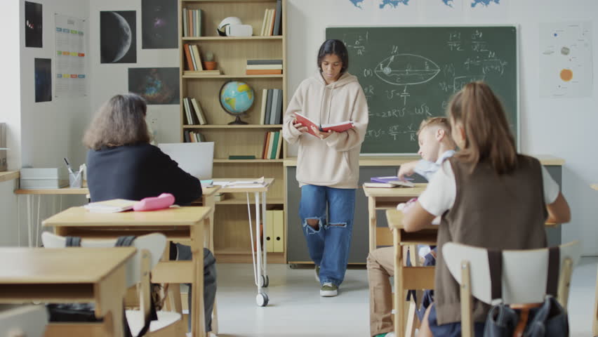 Two teen boys sitting at desks in classroom bullying Black girl making her drop book onto the floor then doing high five and laughing at her | Shutterstock HD Video #1106949349