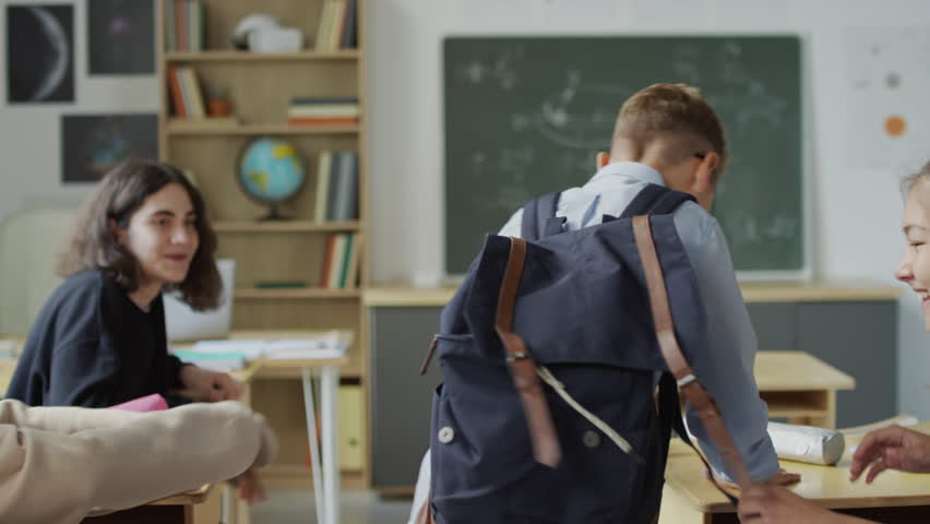 Selective focus shot of teen boy walking along aisle between desks in classroom, his classmates pushing him and trying to | Shutterstock HD Video #1106950457