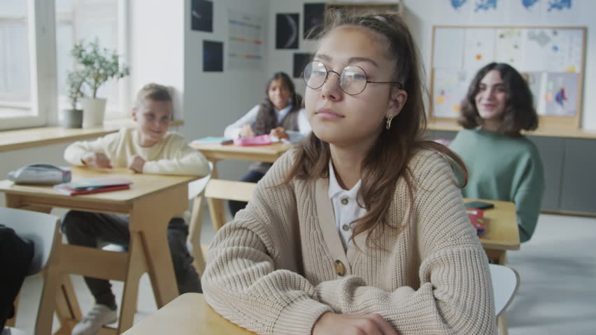 Selective focus shot of depressed teen girl sitting at desk in classroom trying to ignore bullies, who throwing paper balls at her | Shutterstock HD Video #1106954149
