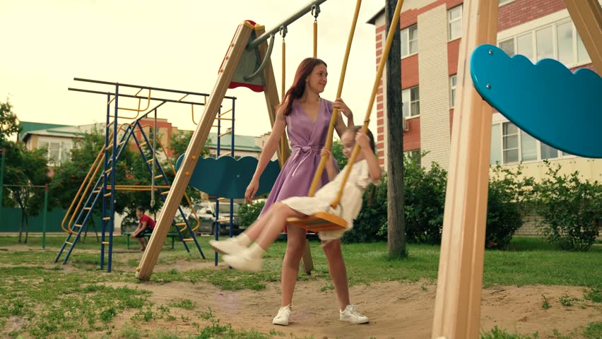 Positive mother and adorable little daughter play swings on playground in park