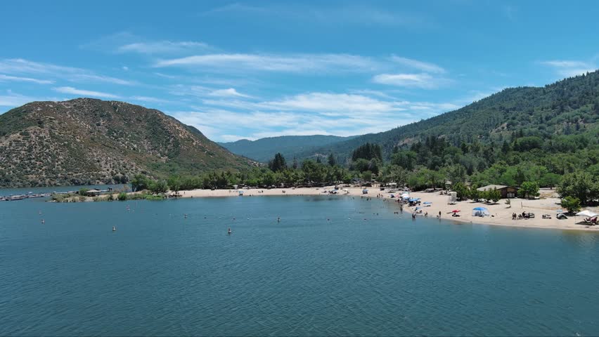 aerial footage of the rippling blue waters of Silverwood Lake with a beach and people swimming in the water and lush green trees, grass and plants, blue sky and clouds in Hesperia California USA