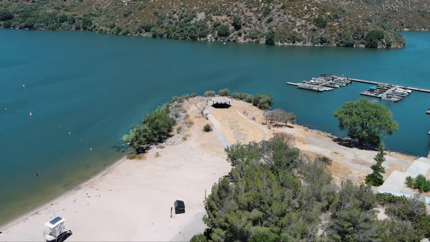 descending aerial footage of the rippling blue waters of Silverwood Lake with a beach, mountains covered in lush green trees, plants and grass, blue sky and clouds in Hesperia California USA
