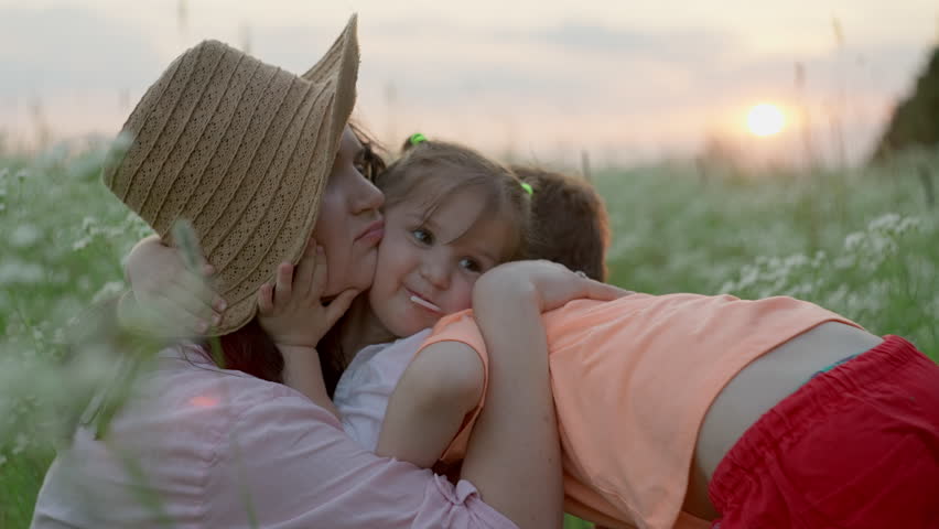 Children hug their mother tightly while sitting on the grass at sunset. Concept of happiness of family and motherly love for son and daughter, smiles and lifestyle. High quality 4k footage Royalty-Free Stock Footage #1106965361