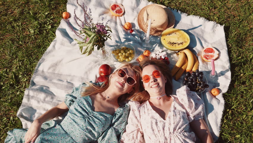 Two young beautiful smiling hipster female in trendy summer sundress and hats. Carefree women making picnic outside. Positive models sitting on plaid on grass, hugging, eating fruits. Top view Royalty-Free Stock Footage #1106966071