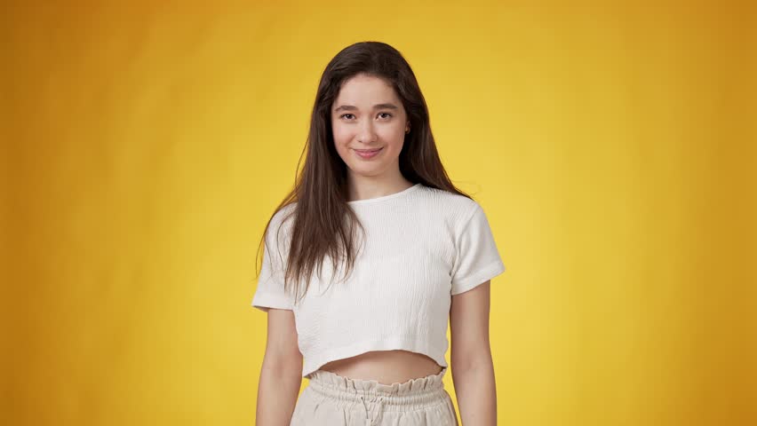Happy casually dressed young smiling woman with long dark hair clenching and shaking her fists at chin level in excitement against yellow background Royalty-Free Stock Footage #1106969353