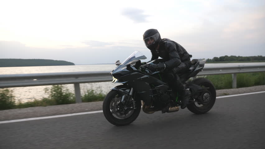 Motorcyclist races his motorcycle on country road near river or lake. Man in helmet rides fast on modern sport motorbike at rural route. Guy driving bike during trip. Concept of freedom and journey