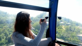 Woman tourist recording video on smartphone sitting in funicular of cable car in mountains. Pretty happy brunette female exploring world travelling in Georgia, Batumi. Travel, tourist, wanderlust.