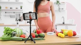Focus on modern cell phone stabilized by tripod on kitchen table among grocery products with lady on screen. Stylish blogger shooting tutorial on developing skills of identifying foods via internet.