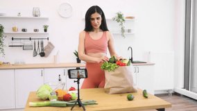 Attractive lady in activewear recording video content about healthy eating using smartphone on tripod in kitchen. Efficient fitness trainer advising about slimming foods on sports and nutrition blog.