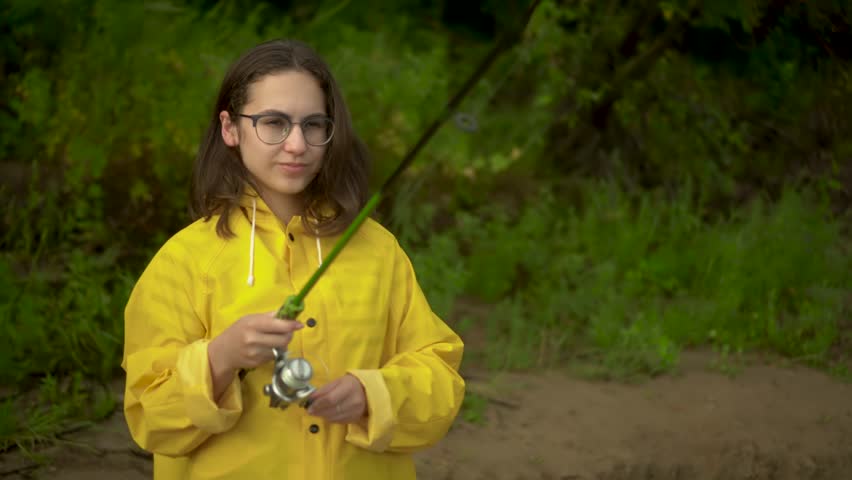 A young woman is fishing on the river bank. A girl in a yellow raincoat with a hood and glasses casts a spinning rod. Close-up.