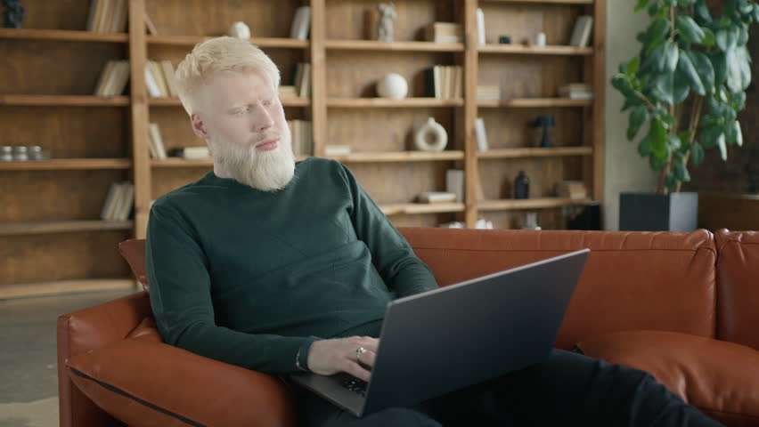 Positive millennial guy freelancer using laptop on sofa at home office living room. Blonde albino entrepreneur touching his beard, working distantly typing on notebook in modern loft apartment 4K slow Royalty-Free Stock Footage #1106982623