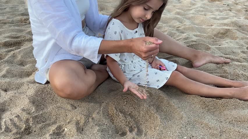 Little girl and her mother having fun playing on the beach sand. Evening sea landscape in the background.