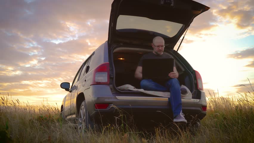 Concentrate confident male freelancer working on laptop, man typing on keyboard, sitting in car trunk while traveling at sunset, general plan. Busy worker freelancer entrepreneur notebook online Royalty-Free Stock Footage #1106984487
