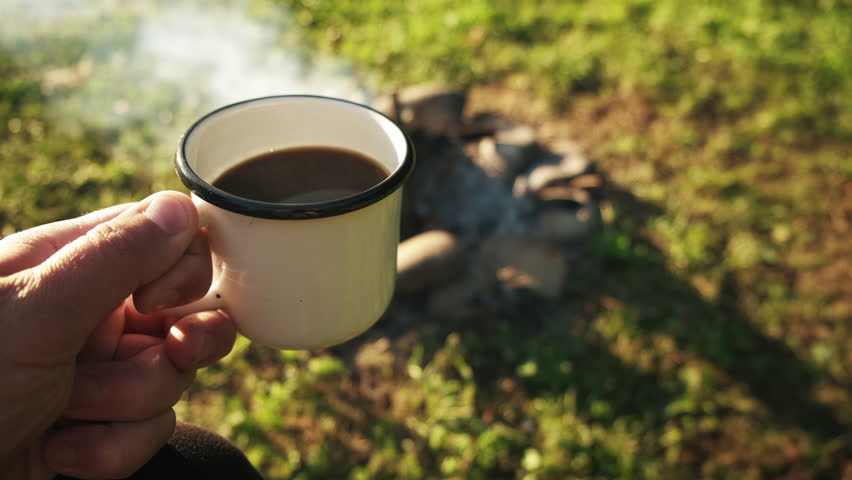 Close up of man traveler hand holding cup near campfire outdoors. Adventure, travel, tourism and camping concept. Hiker drinking coffee from mug at camp. Coffee cooked over bonfire in summer nature