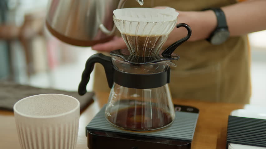 Making Dripped Coffee. Proffesional Barista Pours Hot Water from Drip Kettle Into Coffee Dripper. Aromatic Filtered Coffee Dripping Into Glass Server - Side View