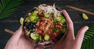 Showing salmon poke bowl, person holding a wooden bowl with raw fish and fresh vegetables, broccoli and edamame beans, high quality video clip, exotic background, palm leaves, 4k footage