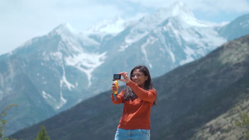 Young Indian woman travel vlogger record video on action camera while standing against snowy mountains in Himachal Pradesh, India. Successful ethnic female following passion. Happy solo tourist enjoy Royalty-Free Stock Footage #1107003399