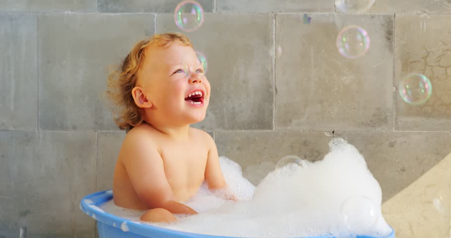 Handsome little boy take bath, sitting in warm water at baby bathtub, slow motion shot. Heap of white fluffy foam on water, large soap bubbles soar down. Short portrait clip of happy child